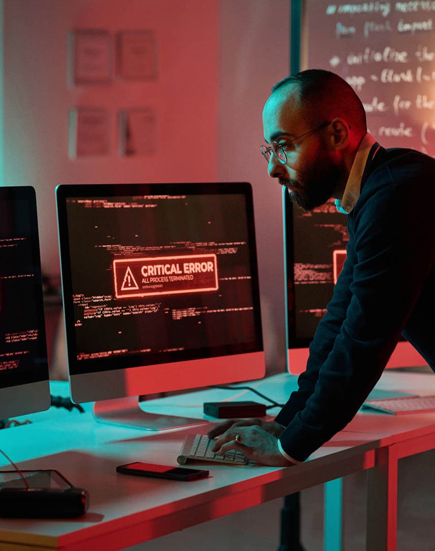 Ein Mitarbeiter vor zwei rot erleuchteten Bildschirmen, auf denen der Hinweis "Critical Error" zu lesen ist