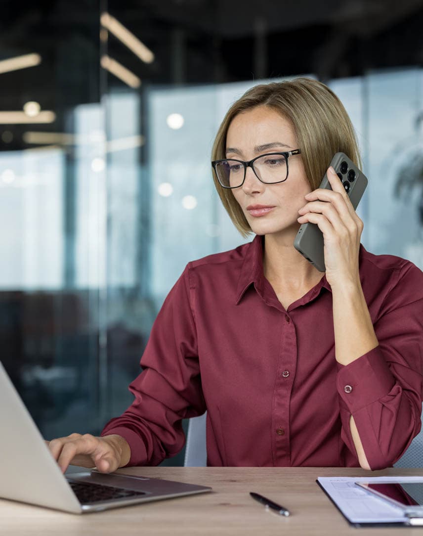 Eine Businessfrau sitzt an einem Schreibtisch mit Laptop und telefoniert mit einem Smartphone.