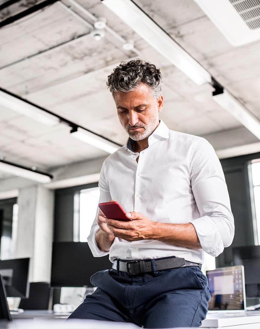 Ein Mann im weißen Hemd sitzt an einen Schreibtisch gelehnt und guckt etwas auf seinem Handy an.