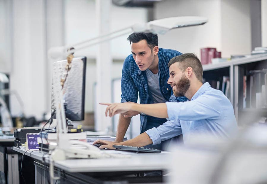 Zwei Männer besprechen im Büro etwas an einem Schreibtisch, der vordere zeigt auf einen Bildschirm.