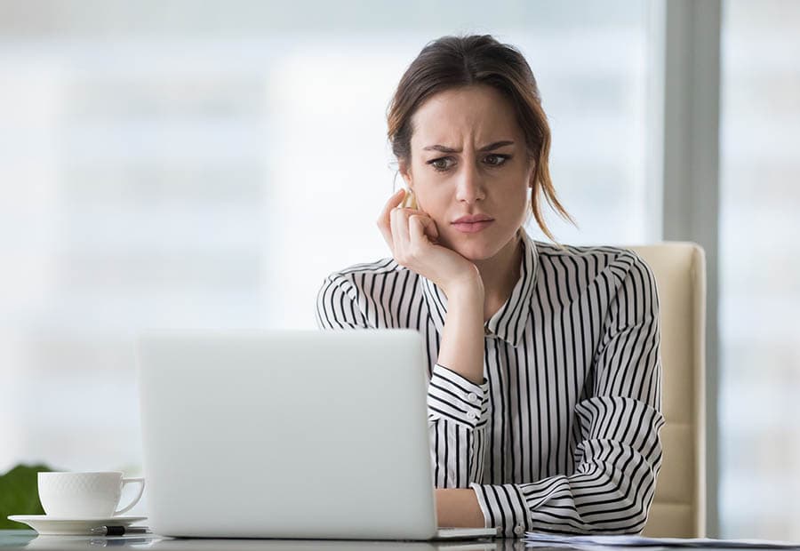 Eine Frau in gestreifter Bluse stützt Ihren Kopf auf Ihre Hand und guckt zweifelnd in einen Laptop auf ihrem Schreibtisch.