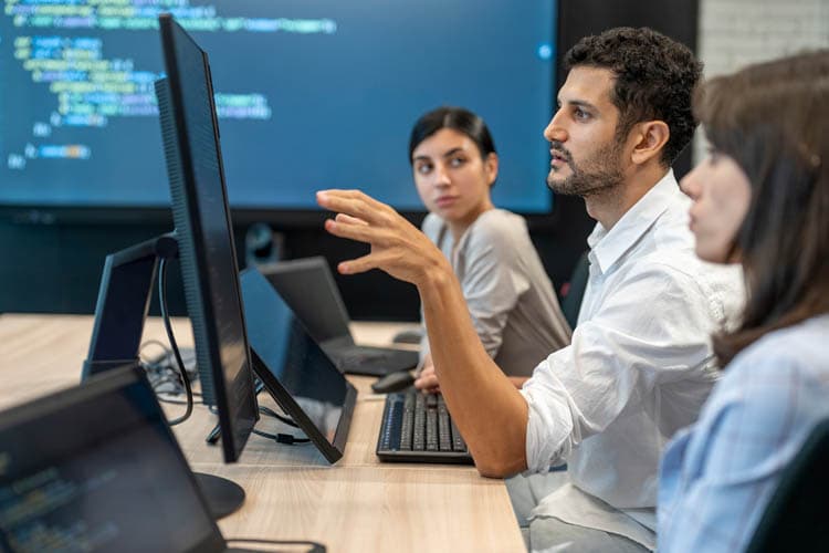 Drei Kolleg*innen vor Computern, im Hintergrund ein großer Screen mit Code