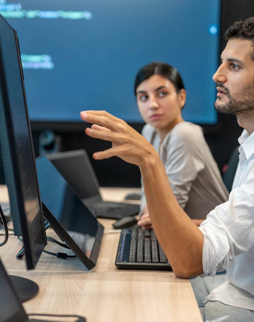 Drei Kolleg*innen vor Computern, im Hintergrund ein großer Screen mit Code
