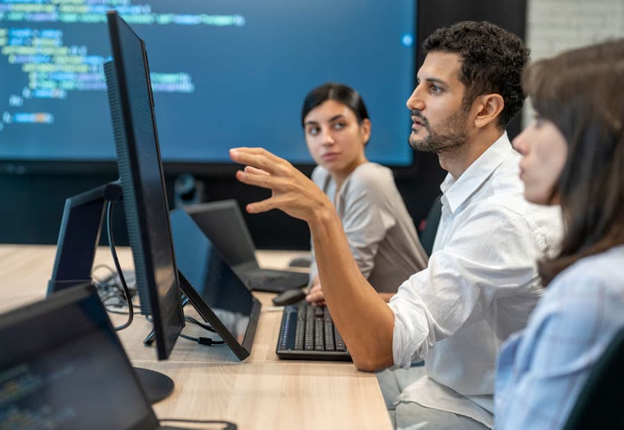 Drei Kolleg*innen vor Computern, im Hintergrund ein großer Screen mit Code