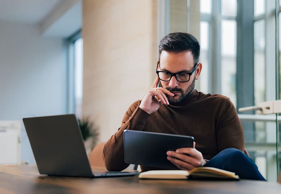 Ein Mann, der an seinem Schreibtisch mit Laptop nachdenkend auf sein Tablet guckt