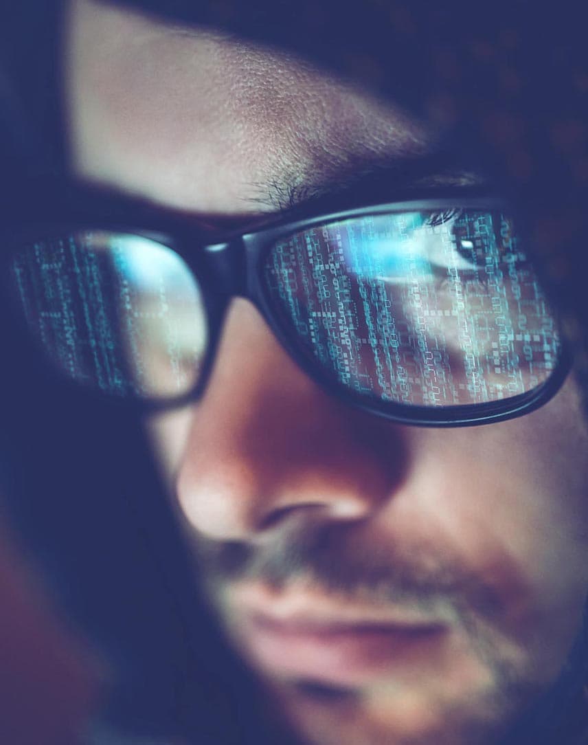 Der Kopf eines jungen Mannes mit Hoodie und Brille, in der sich Computer-Codes spiegeln