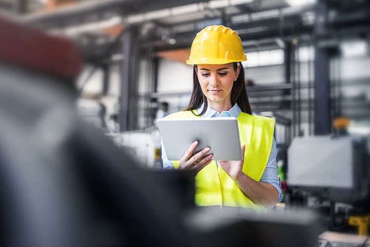 Eine Frau mit Warnweste und Helm arbeitet mit dem Tablet in einer Industriehalle.