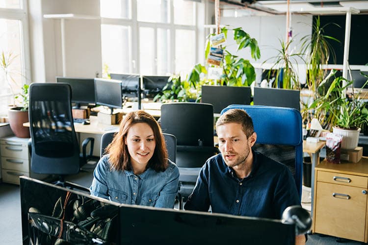 Ein Mann im dunkelblauen Hemd und eine Frau im hellblauen Hemd sitzen vor zwei Bildschirmen in einem Büro.