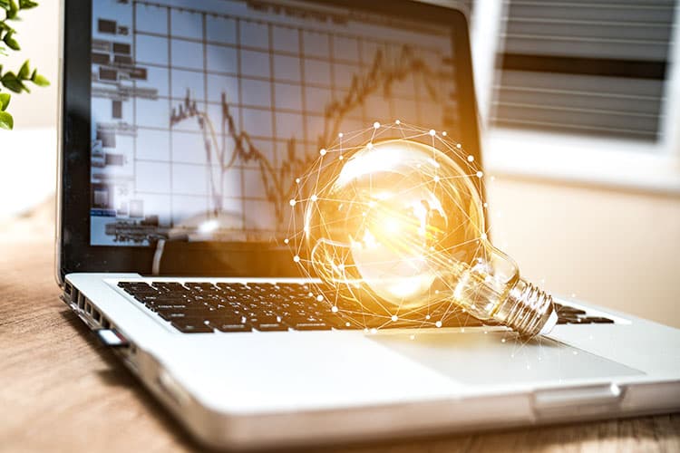 Ein Notebook, auf dem Börsenkurse zu sehen sind und auf dessen Tastatur eine Glühbirne liegt, auf einem Schreibtisch, daneben eine grüne Topfpflanze