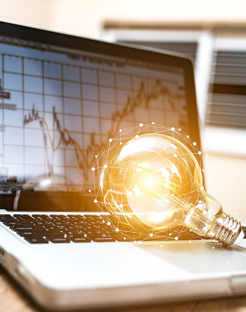 Ein Notebook, auf dem Börsenkurse zu sehen sind und auf dessen Tastatur eine Glühbirne liegt, auf einem Schreibtisch, daneben eine grüne Topfpflanze