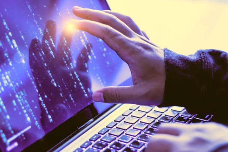 Auf dem Bildschirm eines Laptops sind unleserlich Zeichen und Buchstaben zu erkennen. Eine Hand liegt auf der Tastatur und die andere zeigt auf den Bildschirm. 