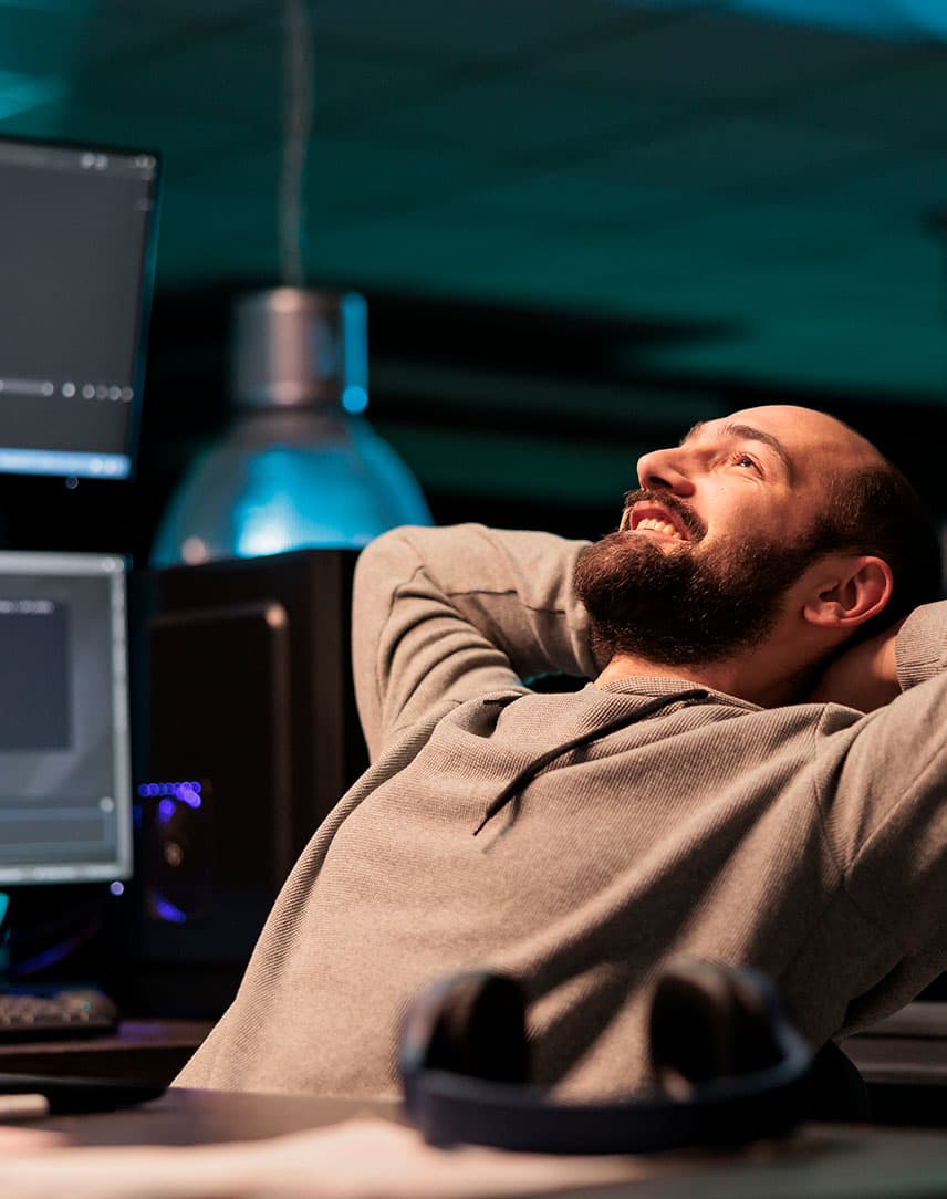 Ein Mann, der am Schreibtisch mit Bildschirmen sitzt, lächelt und die Arme hinter dem Kopf verschränkt