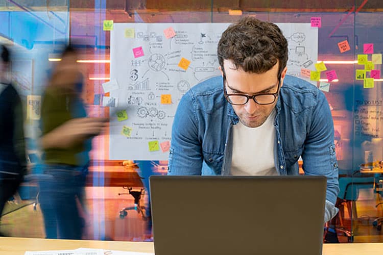 Ein Mann im blauen Hemd, der stehend am Computer arbeitet, im Hintergrund Stellwände mit Post-its sowie Mitarbeitende