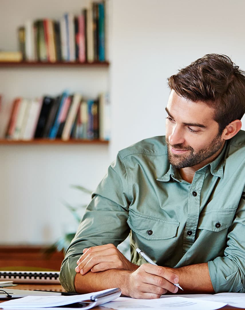 Ein Mann im grünen Hemd sitzt am Schreibtisch, guckt auf seinen Laptop-Bildschirm und macht sich dabei Notizen