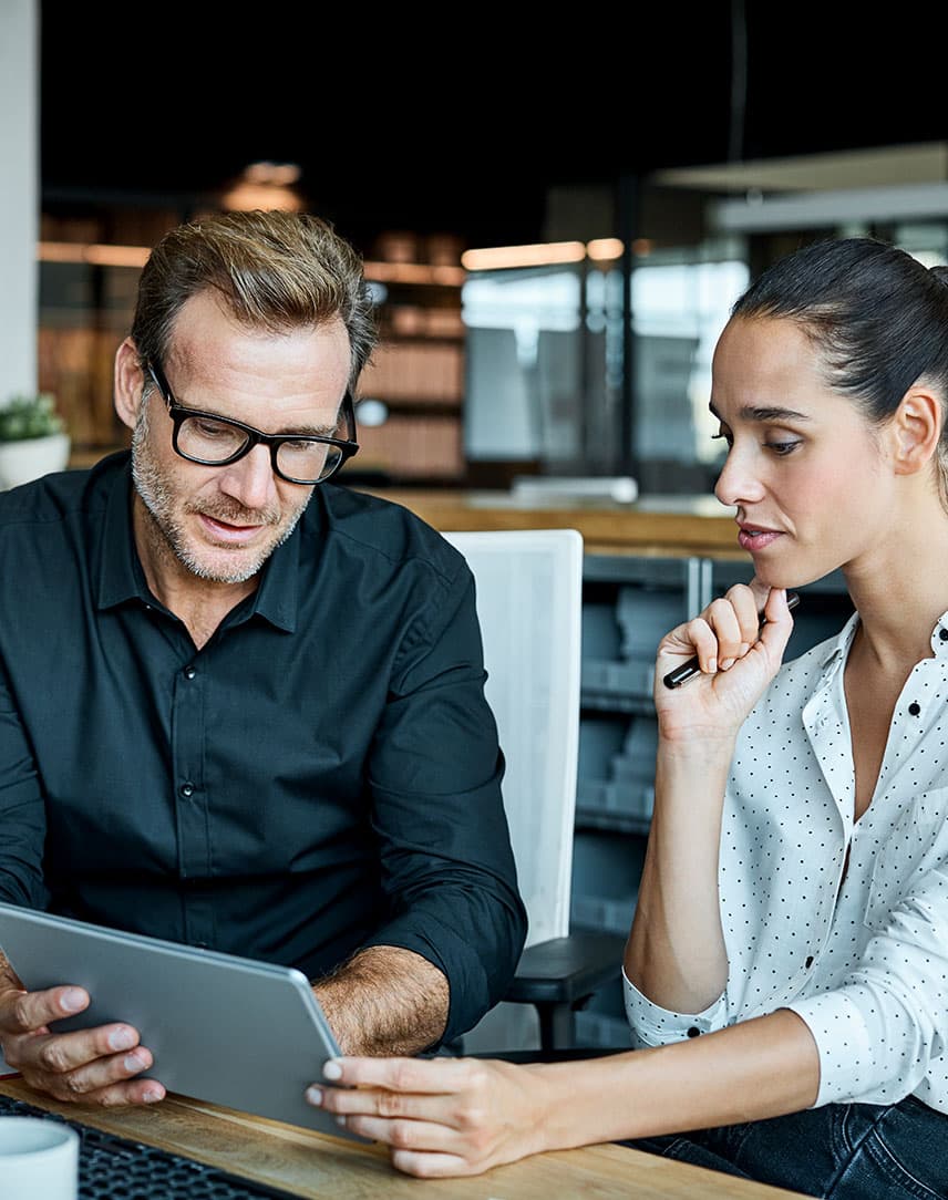 Eine Frau und ein Mann sitzen an einem Schreibtisch in einem Büro und schauen sich etwas auf einem Tablet-PC an. 