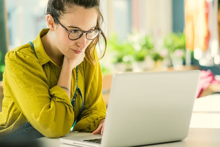 Eine Frau sitzt in ihrem Mode-Atelier an einem Schreibtisch und arbeitet an einem Laptop.