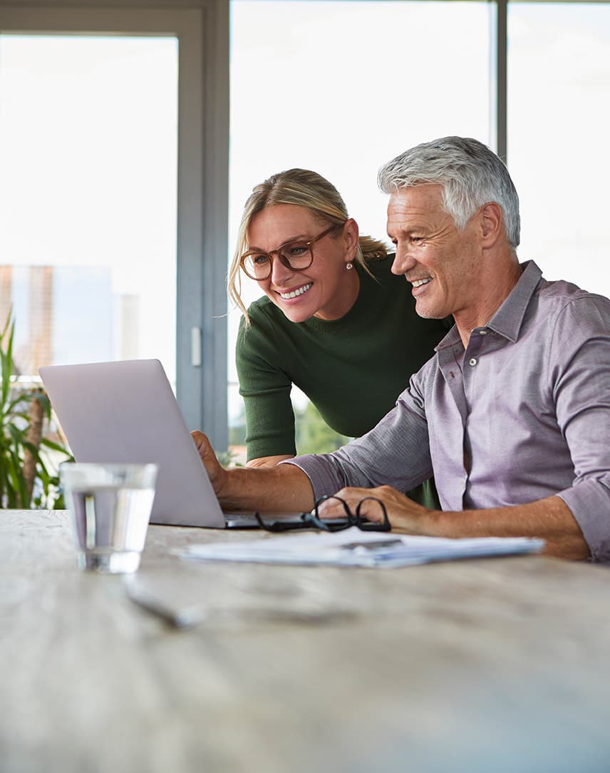 Ein Mann sitz an einem Schreibtisch und arbeitet an einem Laptop. Eine Frau steht neben ihm. Gemeinsam schauen sie auf den Screen. 