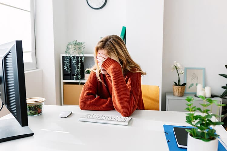 Eine junge Frau sitzt am Schreibtisch und fasst sich nach einer erfolgreichen Phishing-Attacke mit der linken Hand an die Stirn. 