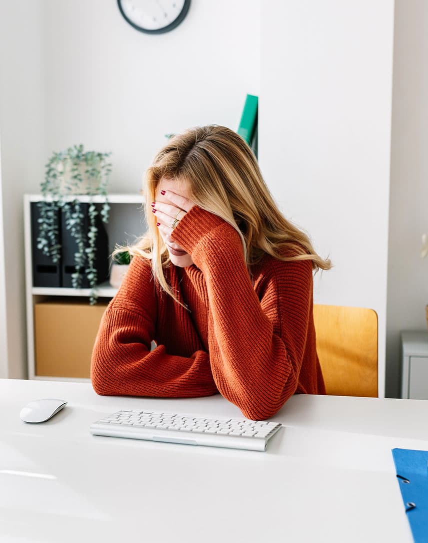 Eine junge Frau sitzt am Schreibtisch und fasst sich nach einer erfolgreichen Phishing-Attacke mit der linken Hand an die Stirn. 