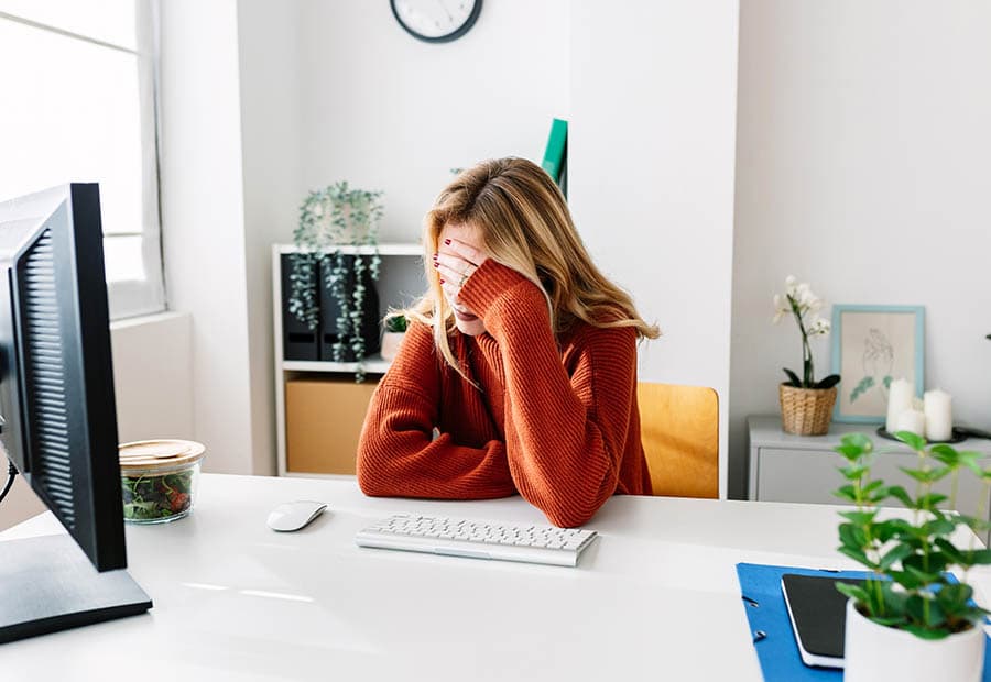 Eine Frau sitzt an einem Schreibtisch vor einem Bildschirm und fasst sich an die Stirn.
