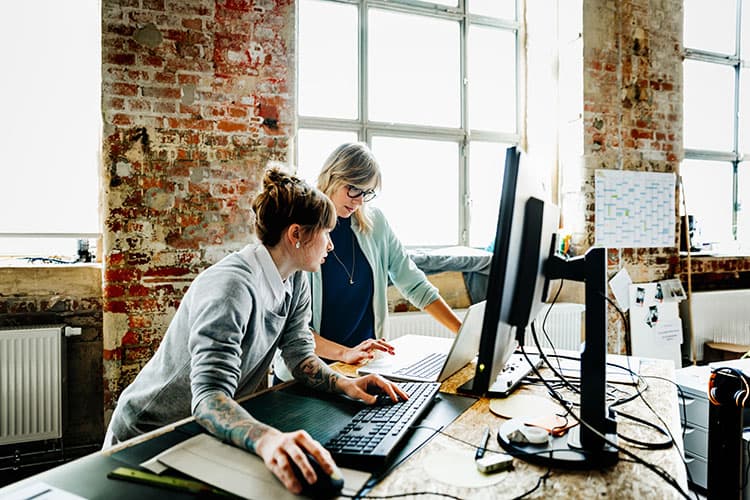 Zwei Frauen stehen an einem Hochtisch und arbeiten an einem Laptop und einem Computer.