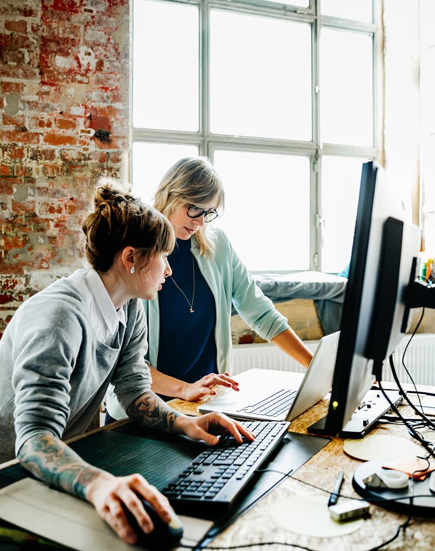 Zwei Frauen stehen an einem Hochtisch und arbeiten an einem Laptop und einem Computer.