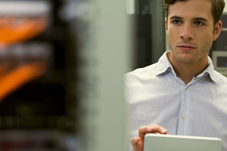 Ein junger Mann steht mit einem Tablet in der Hand in einem Server-Raum eines Rechenzentrums.