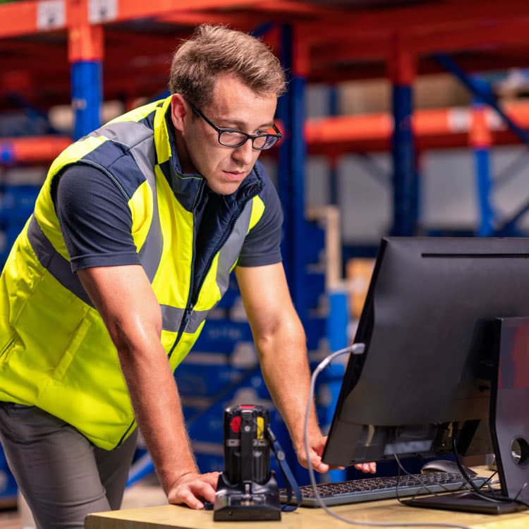 Ein Lagerist mit gelber Warnweste steht im Lager sich auf einen Schreibtisch stützend und guckt auf einen Computerbildschirm.