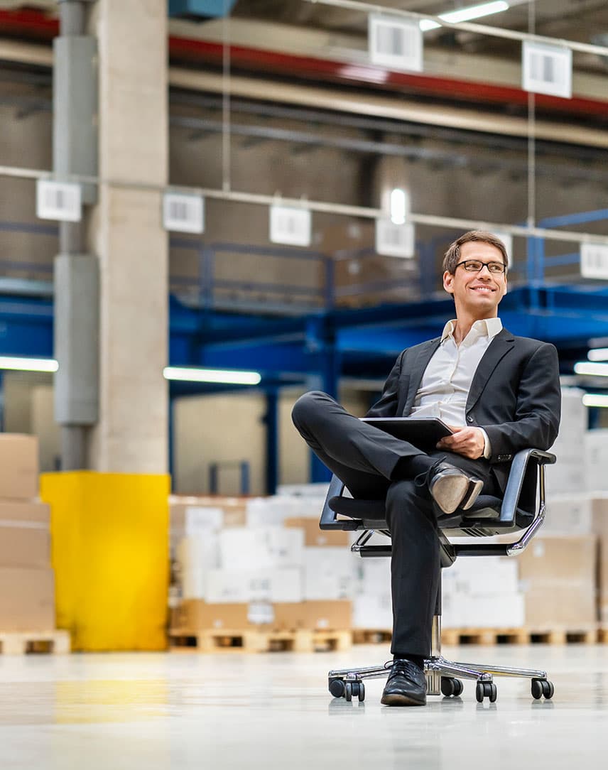 Ein Mann im Anzug sitzt auf einem Bürodrehstuhl im Lager, auf seinem Bein liegt ein Tablet.