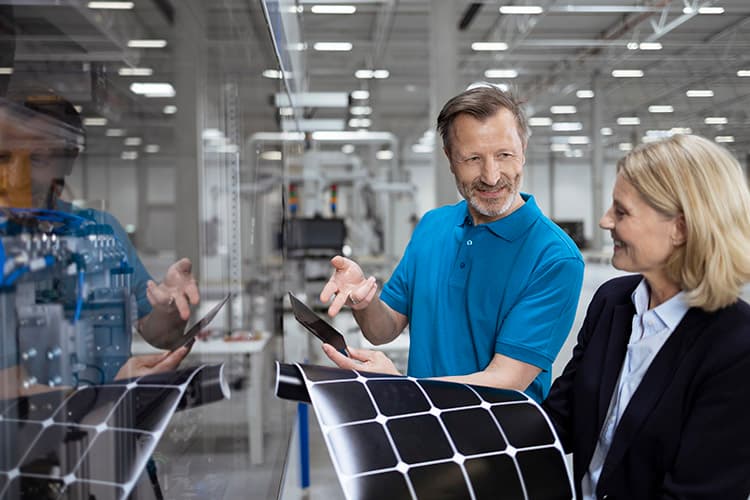 Ein Mann zeigt einer Frau in einem Fabrikgebäude ein Bauteil, die Frau hat eine dehnbare PV-Fläche in den Händen.