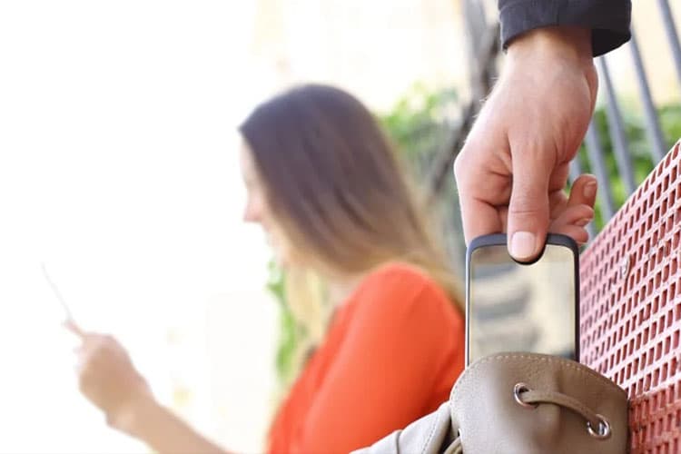 Eine Frau im Hintergrund telefoniert, vorne wird ein Handy aus ihrer Tasche geklaut