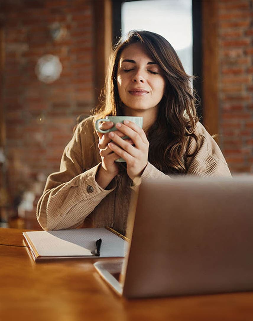 Eine Frau, die mit geschlossenen Augen und Kaffee in beiden Händen an einem Schreibtisch mit Computer sitzt