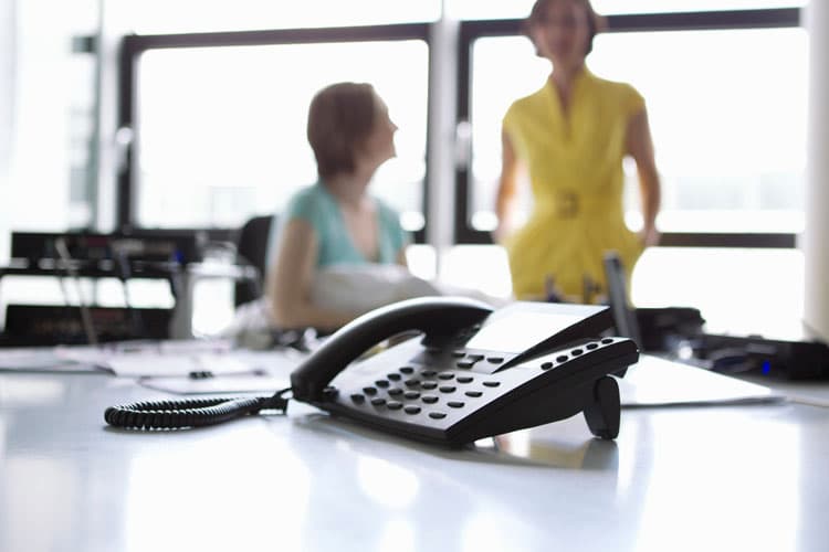 Ein IP-Festnetztelefon, im Hintergrund zwei Kolleginnen