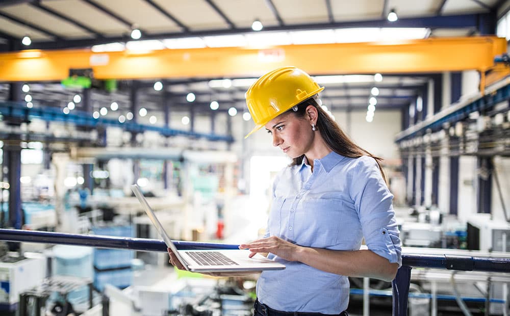 Frau mit Helm in Fabrikhalle schaut auf ihren Laptop.