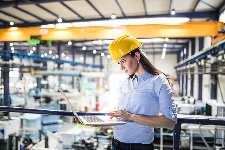 Frau mit Helm in Fabrikhalle schaut auf ihren Laptop.