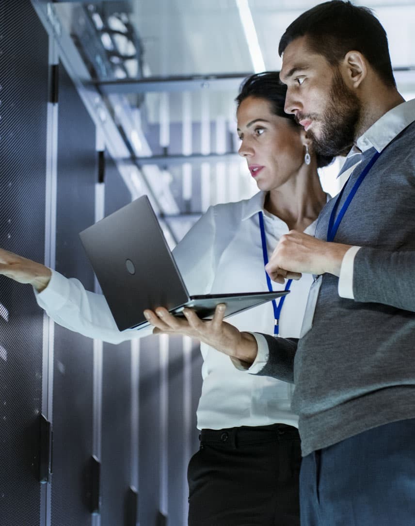 Eine IT-Mitarbeiterin und ein IT-Mitarbeiter prüfen etwas mit Laptop in einem Serverraum.