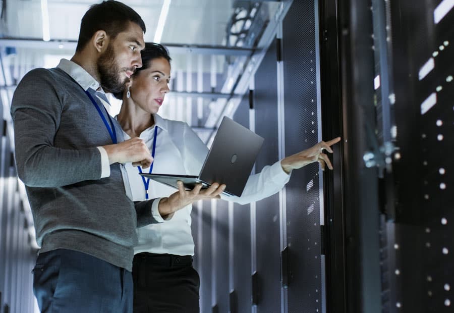 Eine IT-Mitarbeiterin und ein IT-Mitarbeiter prüfen etwas mit Laptop in einem Serverraum.