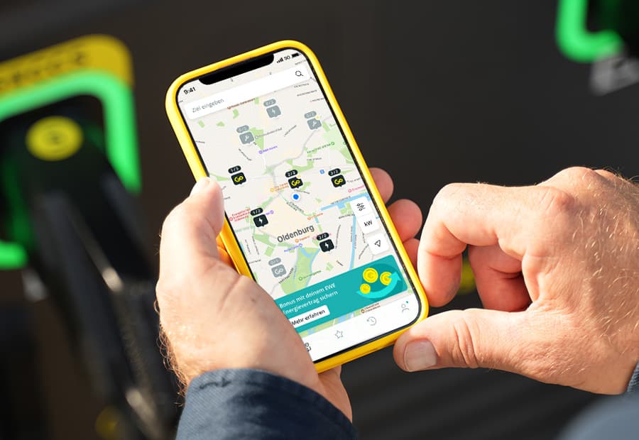 Vor einer EWE GO Ladesäule wird ein Smartphone gehalten auf dem weitere Lade-Standorte zu sehen sind