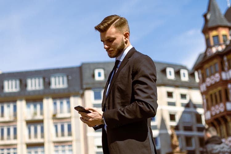 Ein junger Mann im Anzug steht auf einem urbanen Platz und guckt auf seine Smartphone.
