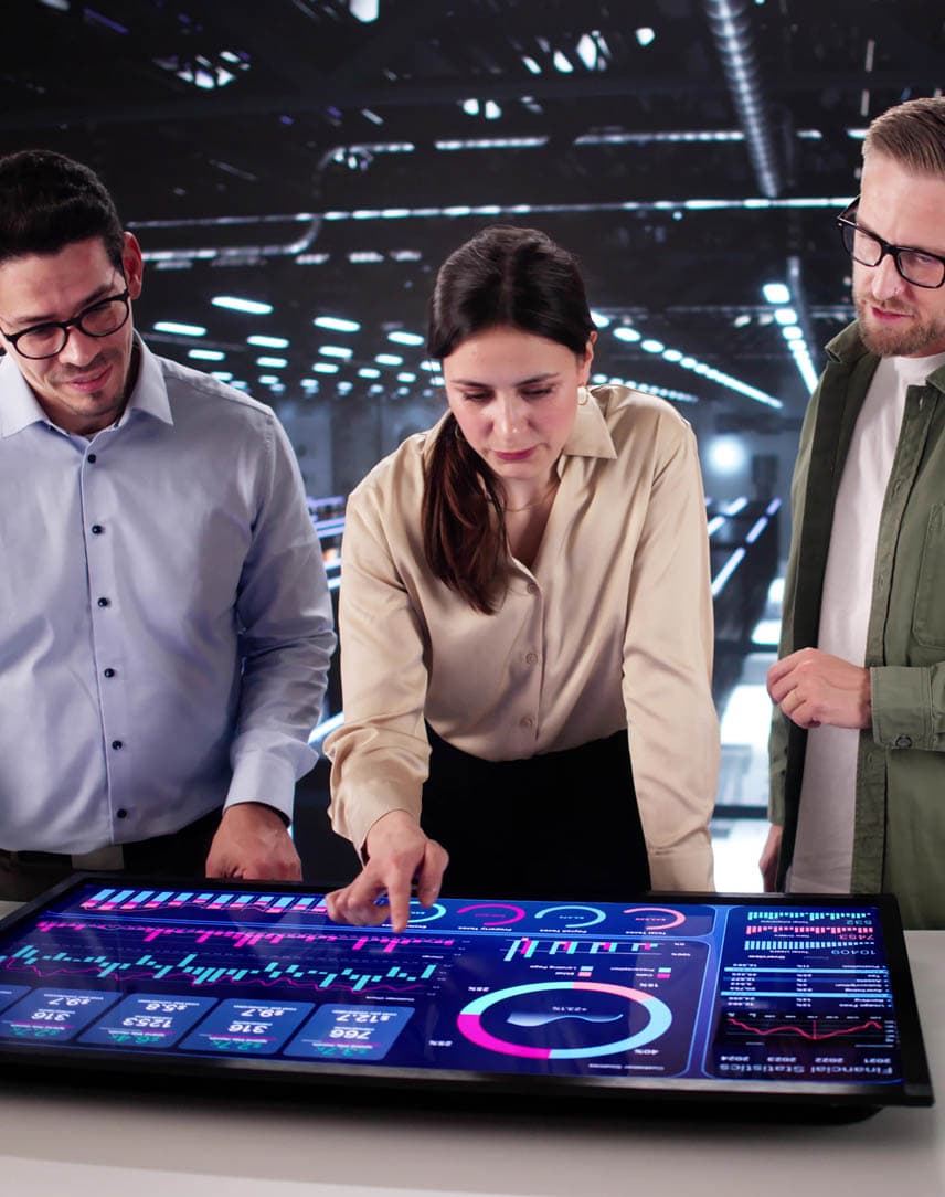 Drei MitarbeiterInnen stehen in einer großen Serverhalle an einem Bildschirm mit Cockpit-Übersicht