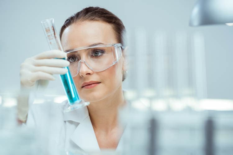 Eine Mitarbeiterin mit Brille und Kittel prüft im Labor ein Reagenzglas.