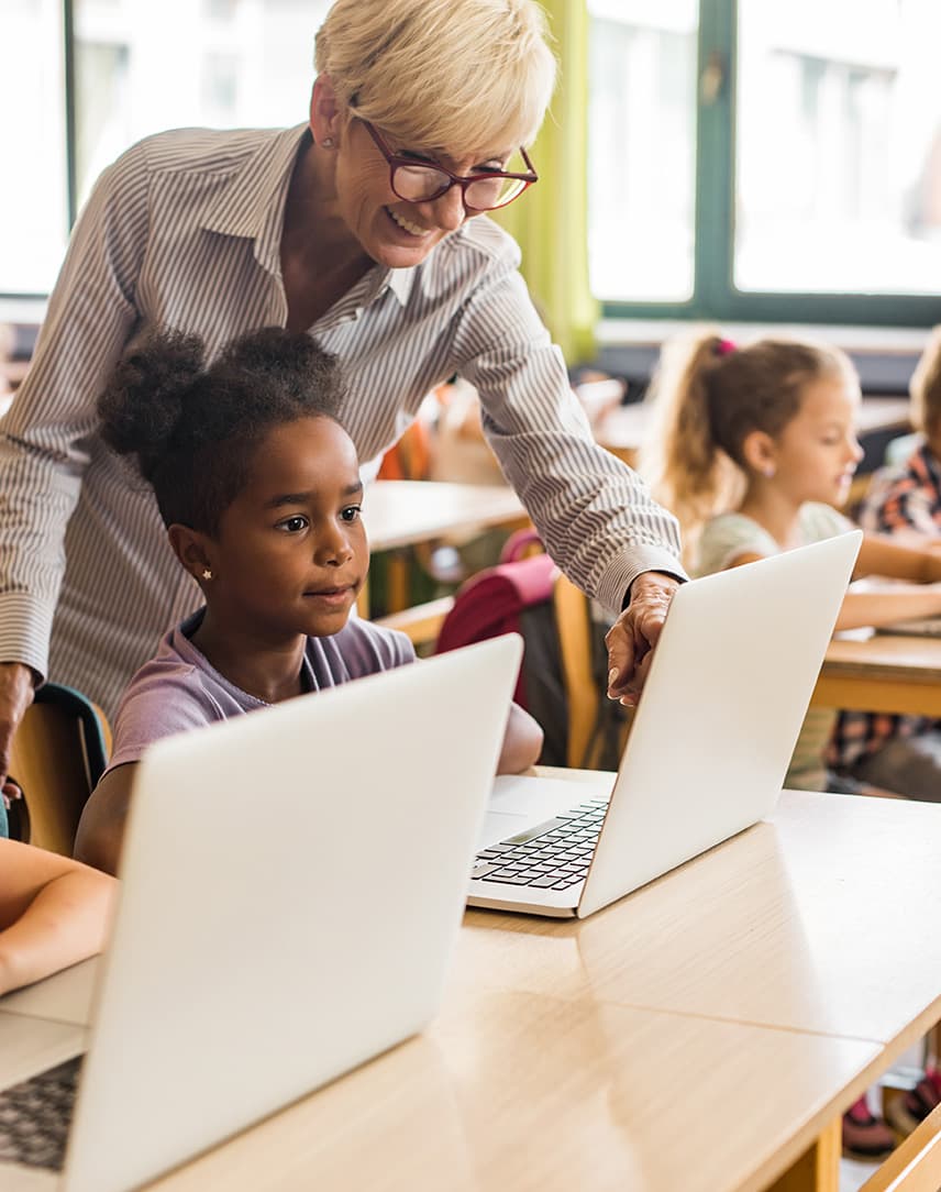 Eine Lehrerin erklärt Schülerinnen und Schülern etwas an Computern im Klassenraum.