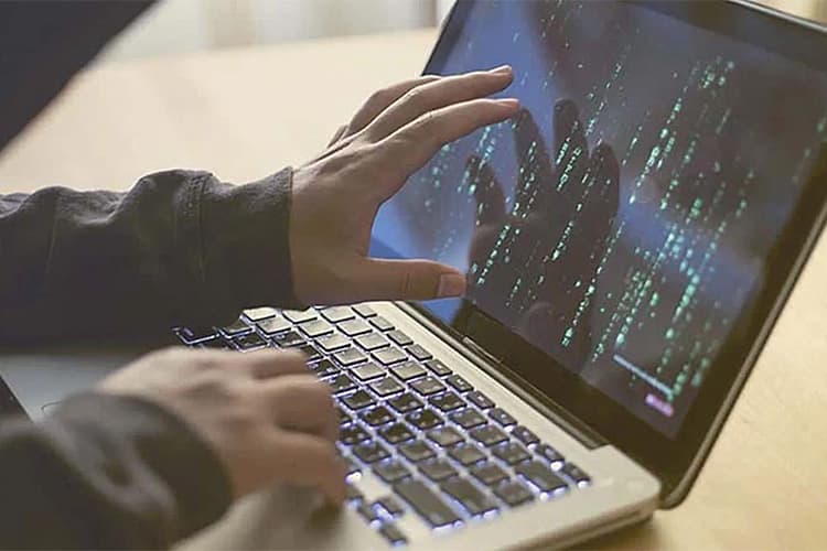 Eine Person im dunklen Hoodie bedient einen Laptop, auf dem binäre Zahlenreihen zu sehen sind.