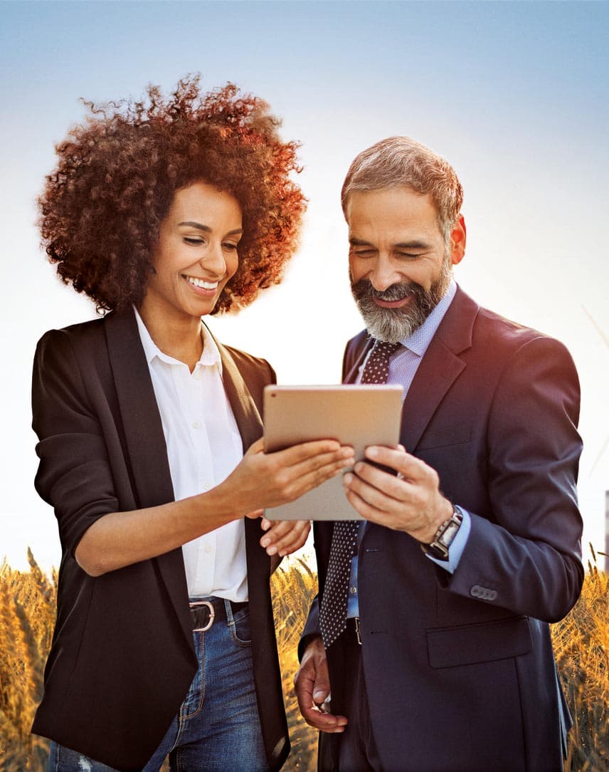 Eine Frau und ein Mann stehen in einem Getreidefeld, sehen auf ein Tablet und lächeln, im Hintergrund ein Fabrikgebäude