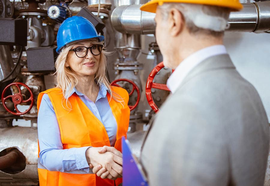 Eine Frau mit Warnweste und Helm schüttelt einem älteren Herren im grauen Sakko mit Helm die Hand, im Hintergrund Rohre einer Kesselanlage