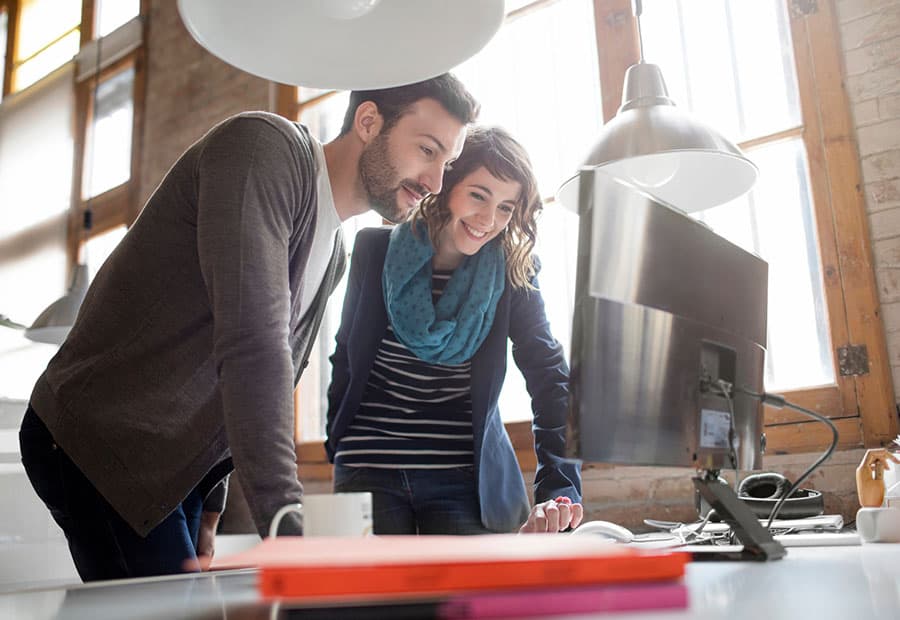 Eine junge Frau und ein junger Mann besprechen in einem Büro stehend etwas an einem Computer auf einem Schreibtisch.