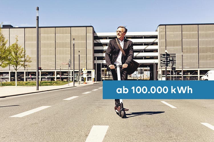 Ein Mann im Anzug fährt mit Tasche auf einem E-Roller auf der Straße aus einem Werksgebäude heraus, darüber ein Hinweis: ab 100.000 kWh