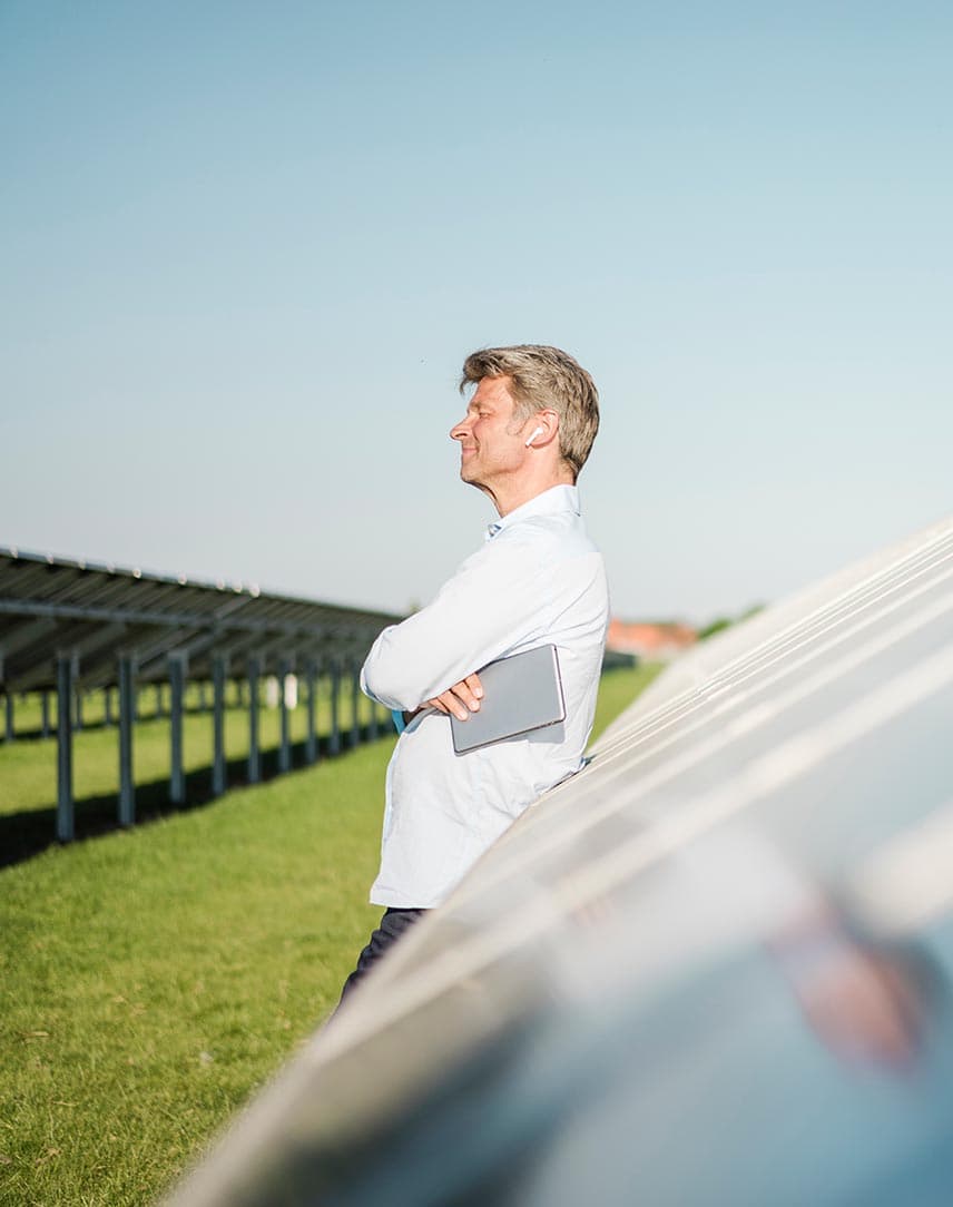 Ein Geschäftsmann lehnt an PV-Modulen auf einer Freifläche und verschränkt die Arme.