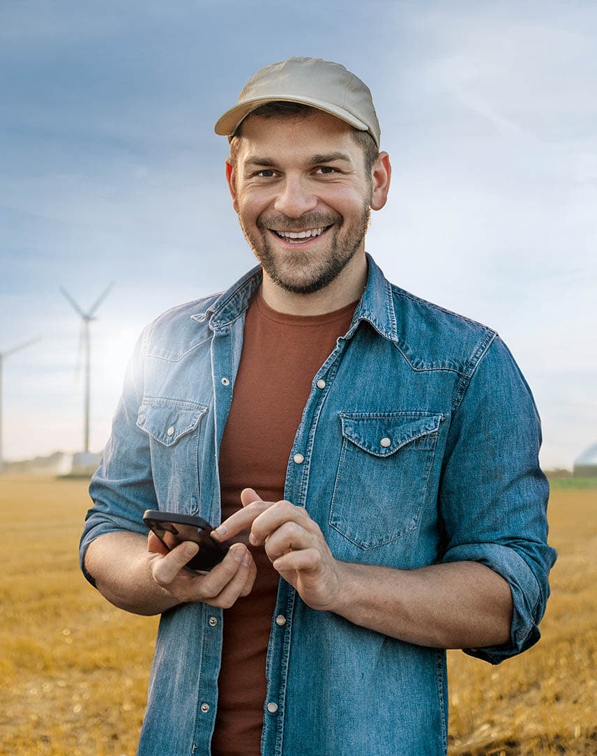 Ein Mann mit Kappe und Jeanshemd, steht auf einem Feld, hält ein Smartphone in der Hand und lächelt, im Hintergrund eine PV-Anlage, Windräder und eine Biogasanlage