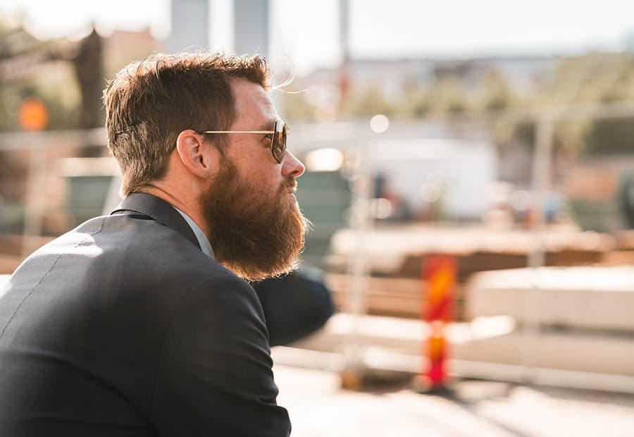Ein Bauleiter im Anzug, mit Bart und Sonnenbrille sieht sich eine Baustelle an.